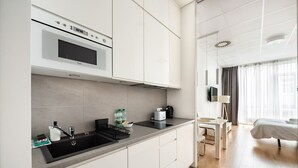 Une cuisine moderne équipée d’un four à micro-ondes encastré, d’un évier et d’un plan de travail sur lequel se trouvent un grille-pain et une bouilloire. À côté de la cuisine se trouve une chambre avec un lit, une table de chevet et un téléviseur.