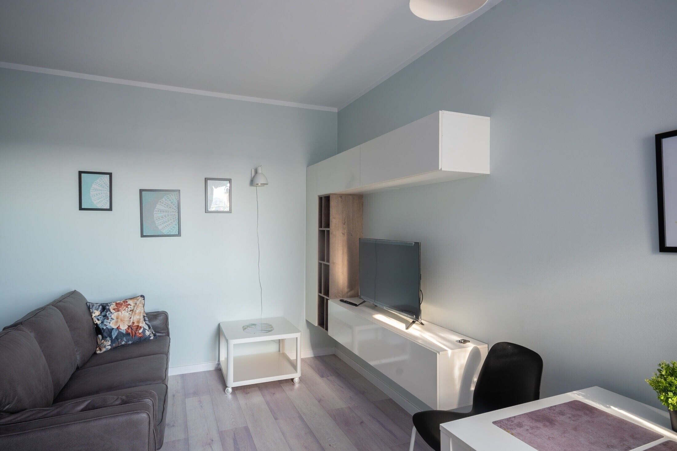 A modern living room with a sofa, a small table, a TV mounted on a white cabinet, and a chair.