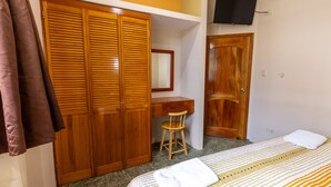A bedroom with a bed, a wooden wardrobe, a desk with a chair, and a television mounted on the wall.