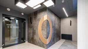 Una entrada moderna a un edificio con una puerta de cristal que indica “miezkania 1-13”, una estructura grande de madera y hormigón, y un radiador en el suelo.