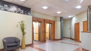 Un hall d’entrée avec un canapé gris, un grand vase rempli de fleurs et des portes en bois donnant accès aux ascenseurs.