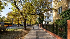 A tree-lined sidewalk with a brick wall and metal fence on one side, and a building with a balcony on the other. - Saska Kępa Loft Style Apartment (Warszawa)