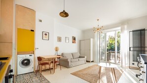 A modern living room with a sofa, dining table, and chairs. There is a washing machine in the corner, a rug on the floor, and a large window with a view of the outdoors. - Saska Kępa Loft Style Apartment (Warszawa)