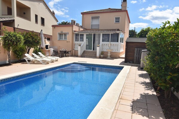 A swimming pool with lounge chairs, a covered patio, and a two-story building with a balcony. - Charming House in Roses, Costa Brava: Private Pool, Close to the Beach, Ideal for Families and Groups. (Roses)