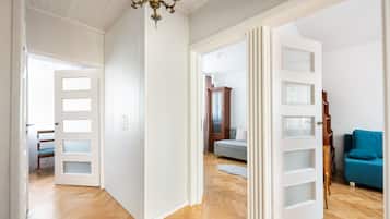 Ein Flur mit Holzboden, weißen Wänden und einer Chandelierleuchtre. Eine Tür führt in einen Raum mit einem Bett und einem blauen Sessel.