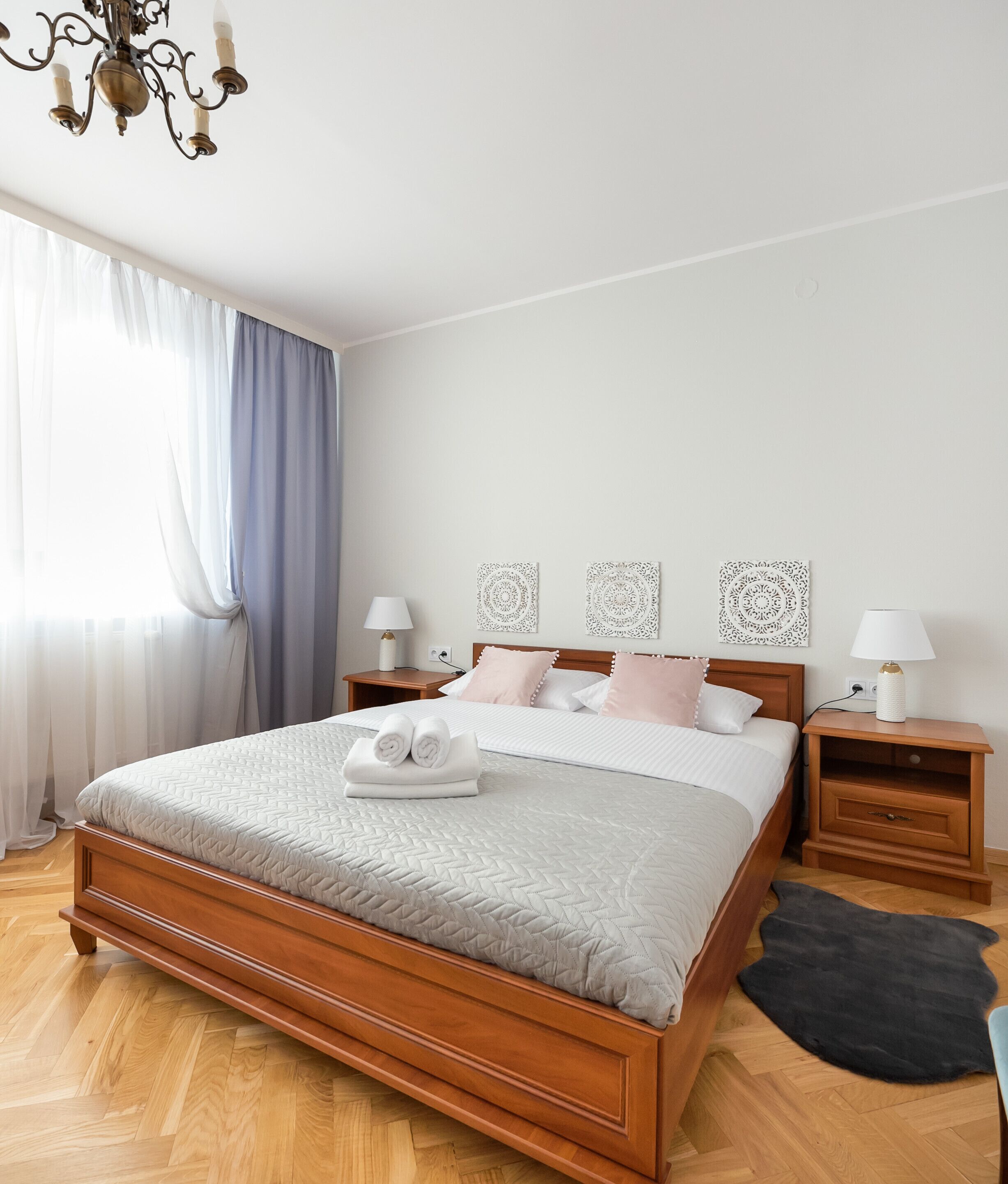 Een slaapkamer met een houten bed, twee nachtkastjes met lampen, een kroonluchter en een groot raam met gordijnen.