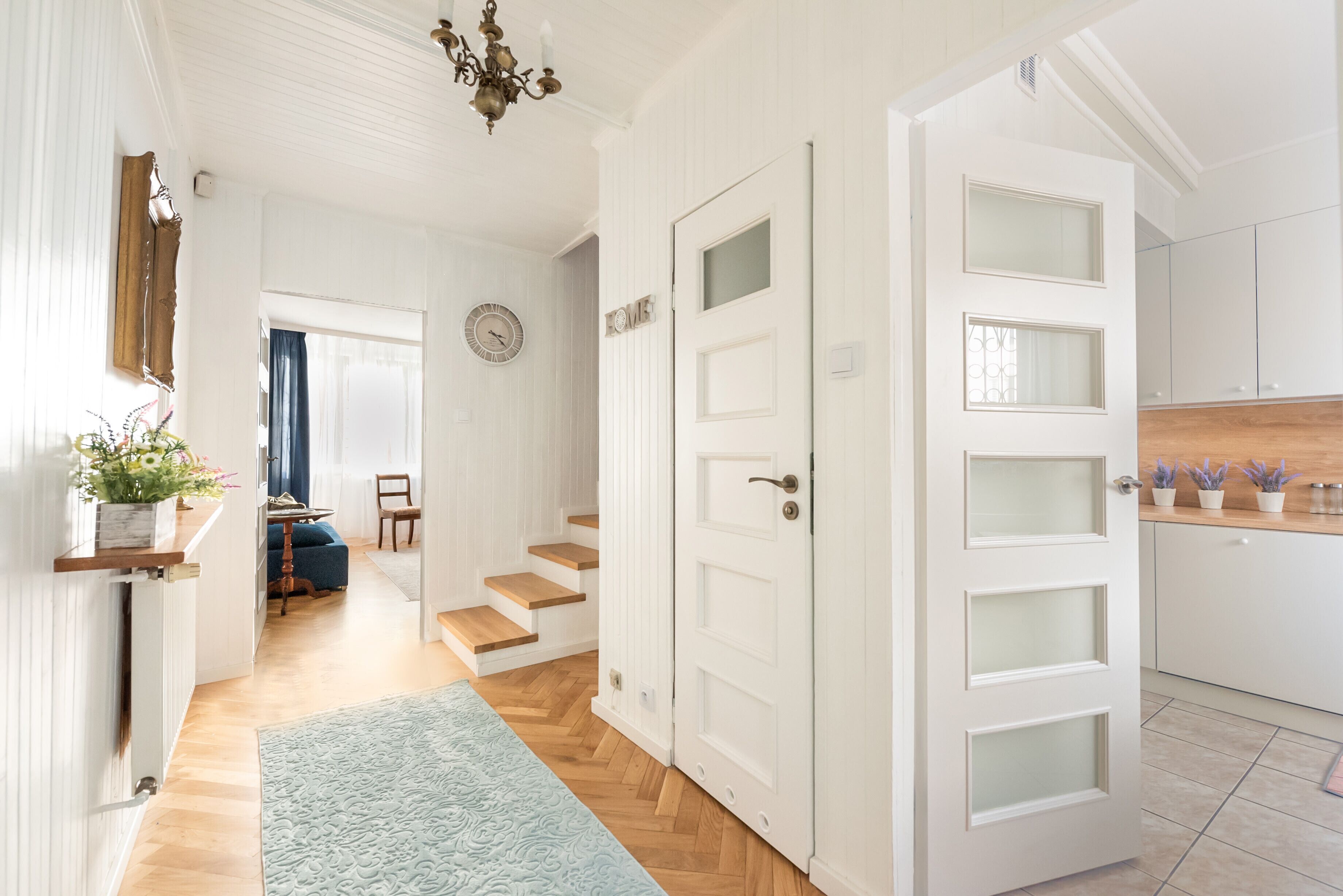 Ein Flur mit Holzboden, einem blauen Teppich, einer weißen Tür mit geätzten Glasscheiben und einer Treppe, die in ein weiteres Zimmer führt.