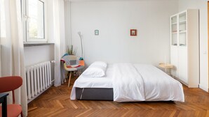 Un dormitorio con cama, una silla, un radiador, una ventana y una lámpara en el techo.