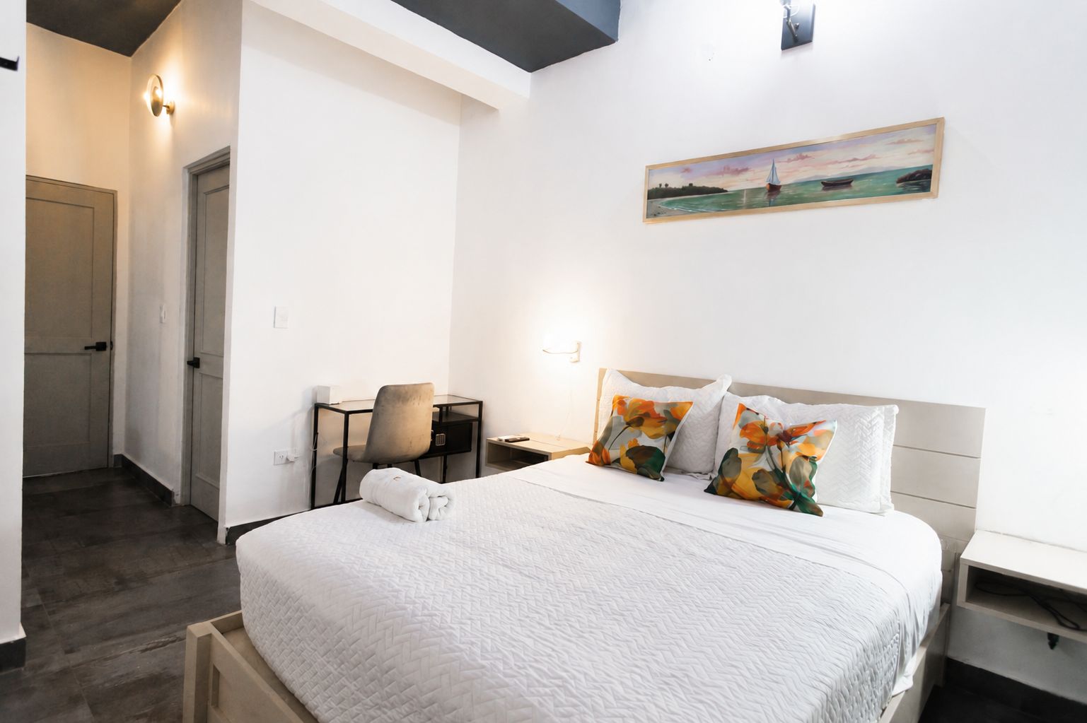 A hotel room with a bed, a desk, a chair, a painting, and a door.