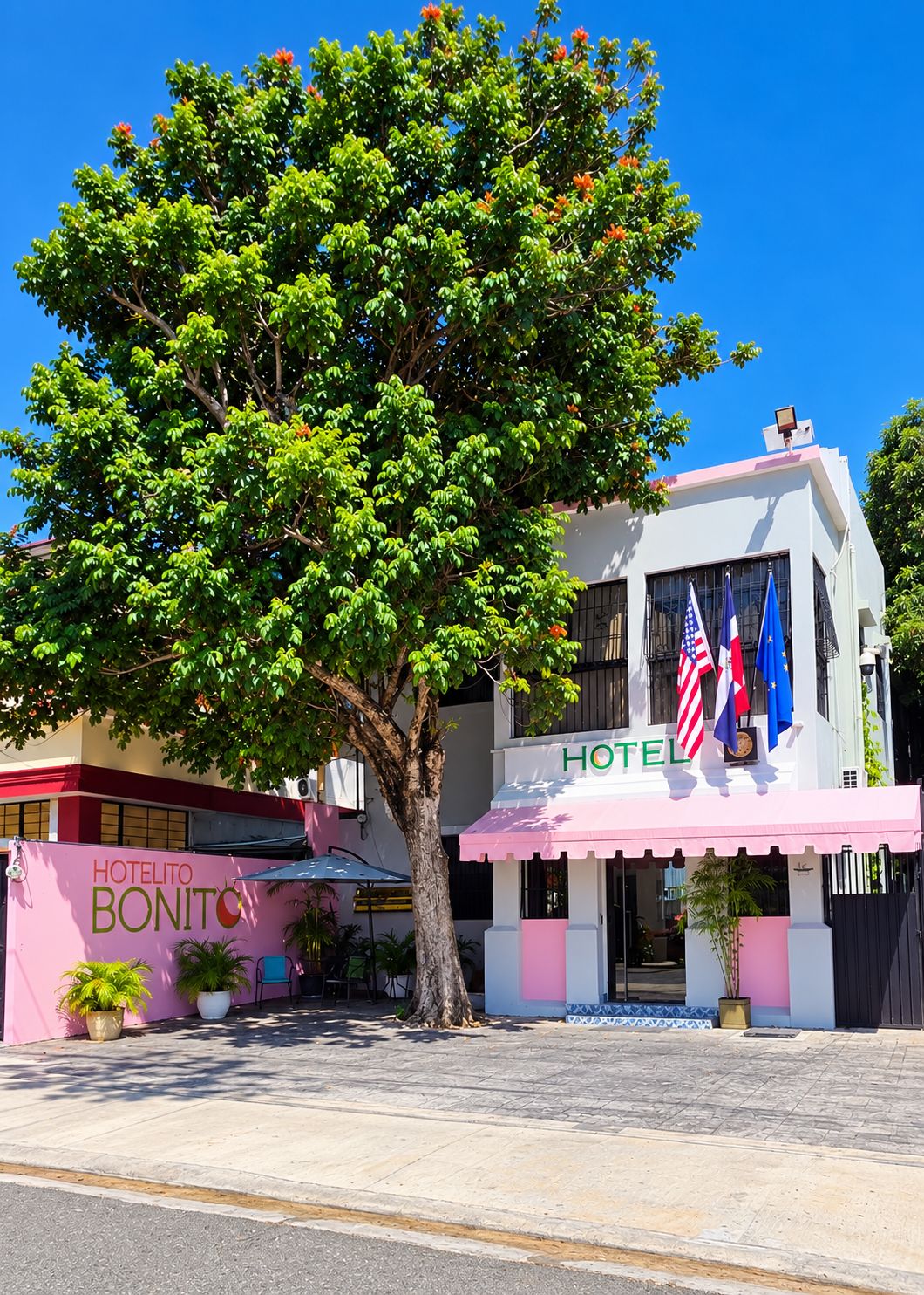 Un hôtel avec un auvent rose, des drapeaux et un grand arbre devant.