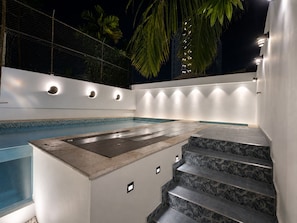 Un área de piscina moderna al aire libre con una bañera de hidromasaje integrada, una escalera con diseño de hojas tropicales e iluminación ambiental.