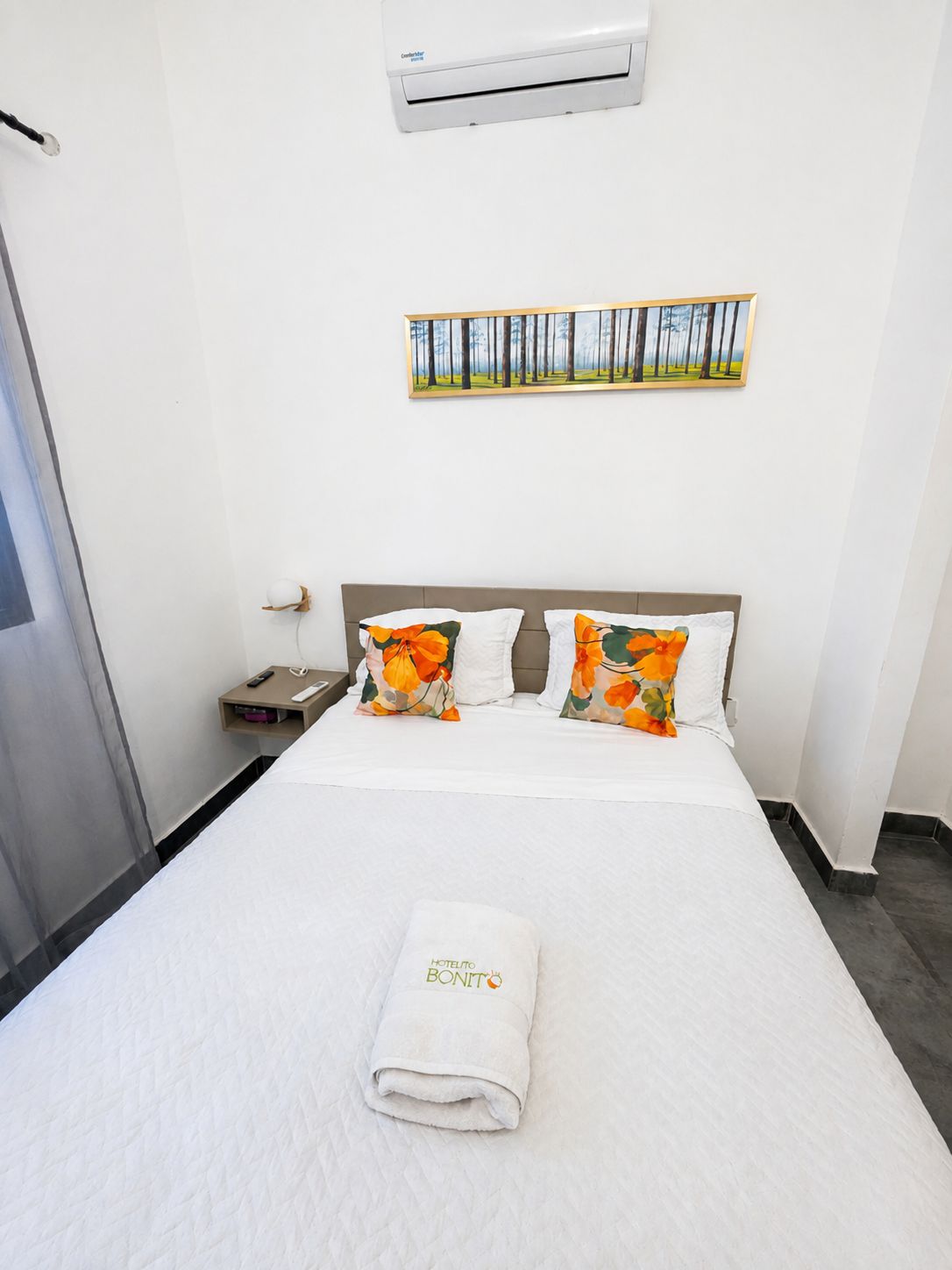 A hotel room with a bed, a painting on the wall, and an air conditioning unit.