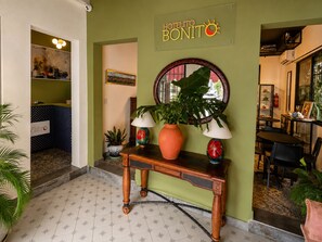 A hotel lobby with a reception desk, a large potted plant, a wall-mounted sign, and a dining area with chairs and tables.
