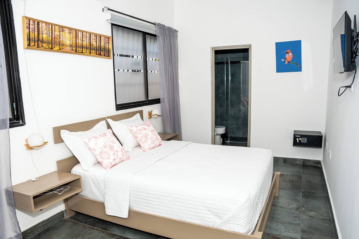 Een slaapkamer met een bed, nachtkastjes, een raam met gordijnen, een schilderij, een douche en een aan de muur bevestigde televisie.