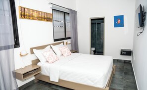A bedroom with a bed, bedside tables, a window with curtains, a painting, a shower area, and a wall-mounted TV. - Hotelito Bonito Eli & Edw (Santo Domingo)