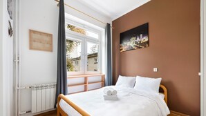 Un dormitorio con cama de madera, ropa de cama blanca, vista por una ventana y un cuadro de paisaje urbano en la pared.