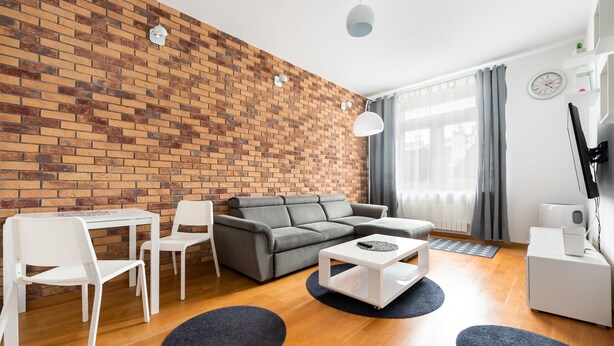 Una sala moderna con pared de ladrillo, sofá gris, sillas blancas, una mesa de centro y un televisor sobre un soporte blanco.