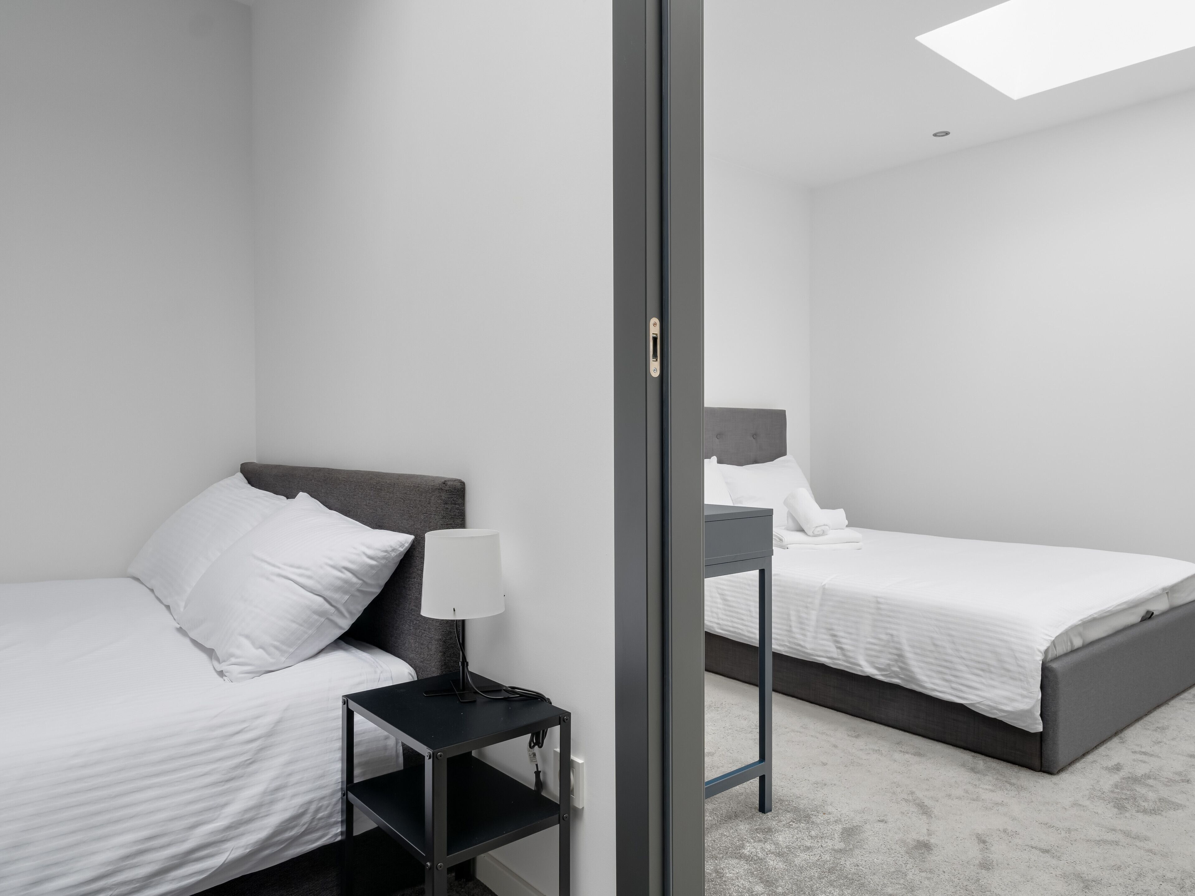 Una habitación de hotel moderna con dos camas, una mesita de noche con lámpara y vista a la ciudad a través de un ventanal.
