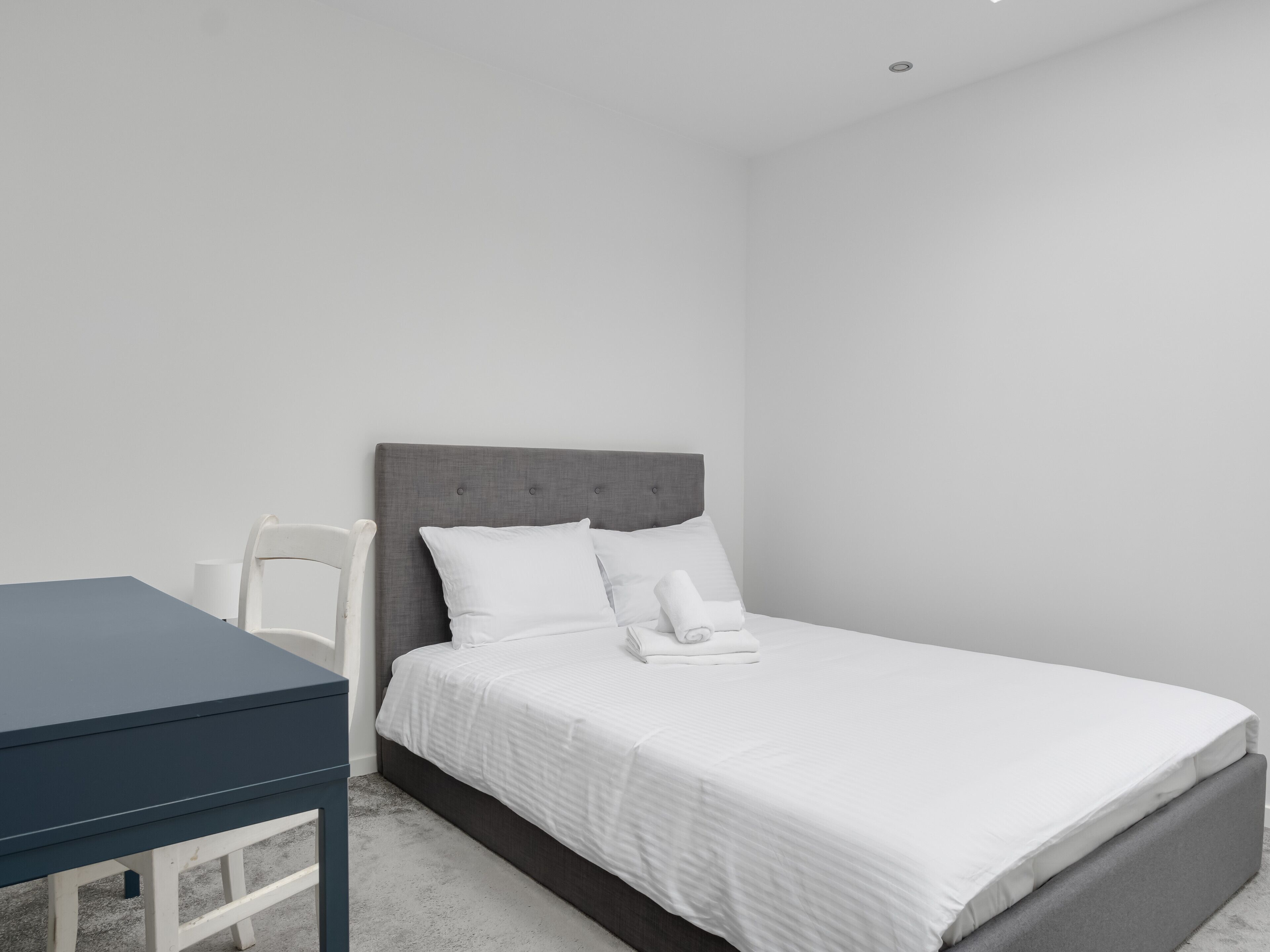 A neatly made bed with white bedding, a grey upholstered headboard, and a white chair beside it.