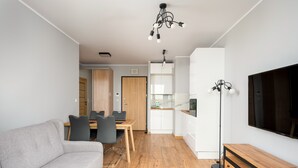 A modern living room with a grey sofa, wooden flooring, a dining area with chairs, a kitchen with white cabinets, and a TV on the wall. - Wiktoryn Comfortable Two-Room Apartment (Warszawa)