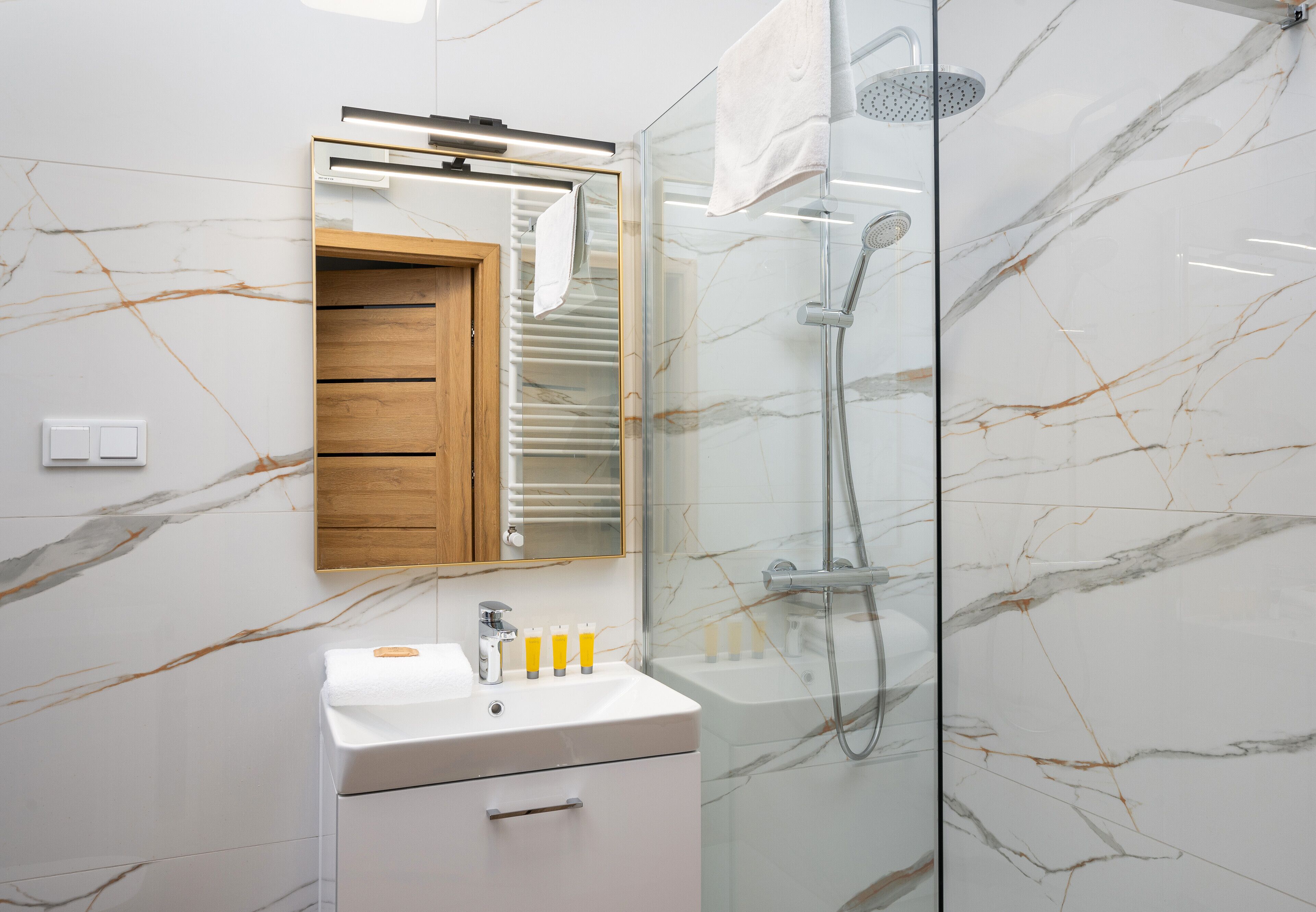 Une salle de bain moderne avec un mur en marbre, un meuble à pharmacie en bois, un lavabo blanc avec des articles de toilette et une douche séparée par une paroi en verre.