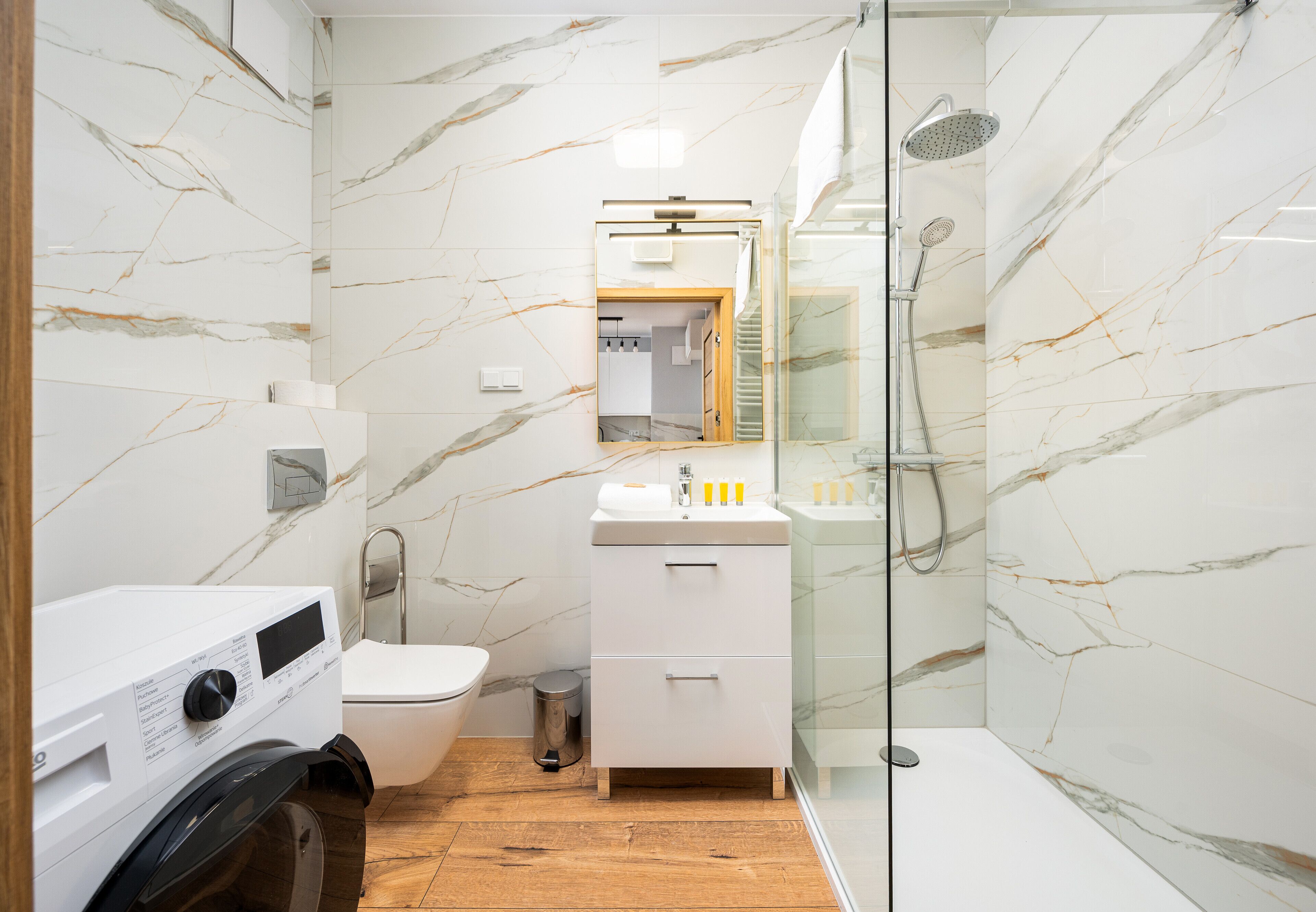 Un baño moderno con salpicadero de mármol, cabina de ducha acristalada, lavabo blanco y lavadora.