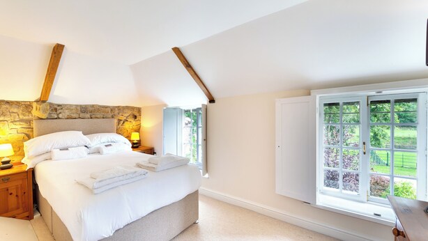 Une chambre avec un mur en pierre, un grand lit, une table de chevet avec une lampe et une fenêtre donnant sur un paysage verdoyant.