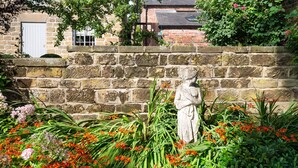 Un mur de pierre orné d’une statue, dans un jardin où poussent différentes sortes de fleurs.