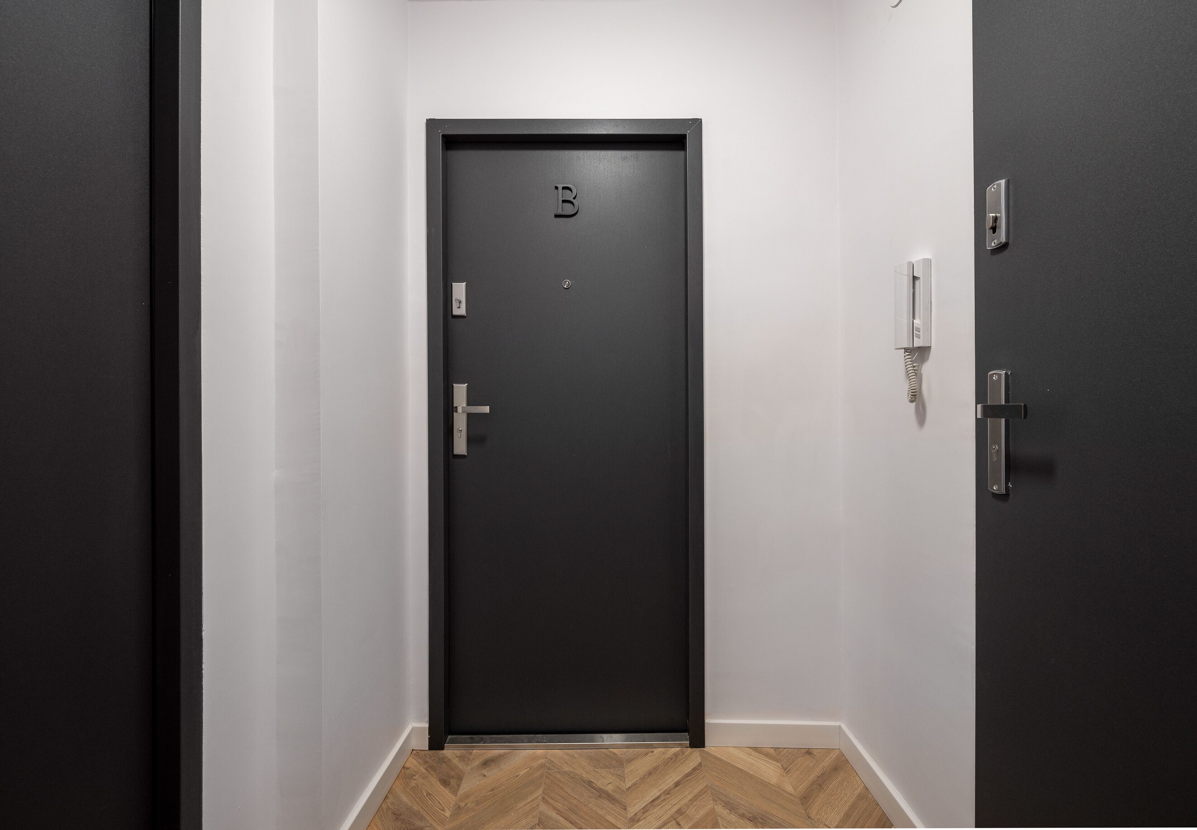 A dark-colored door with a letter 'B' on it, a doorbell, and a wall-mounted intercom.