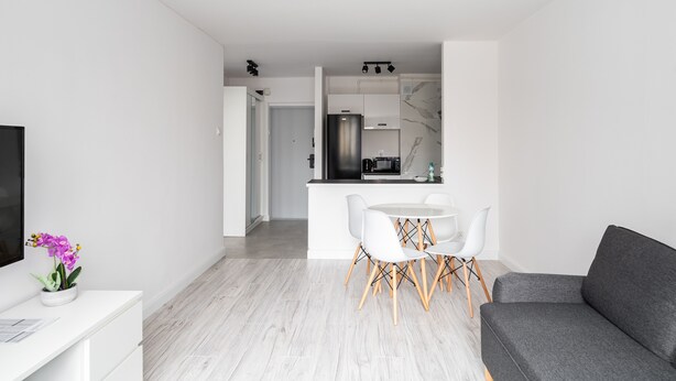 Una sala de estar moderna con un sofá gris, una mesa de comedor blanca con sillas, y una cocina al fondo.