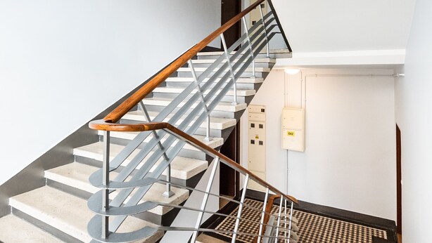 A modern staircase with a wooden handrail and metal balusters, leading to a landing with a light switch and a door. - Old Town 5-Bedroom Apartment (Warszawa)