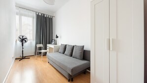 A modern, minimalistic living room with a grey sofa, a small white desk, a standing fan, and a white wardrobe. - Old Town 5-Bedroom Apartment (Warszawa)
