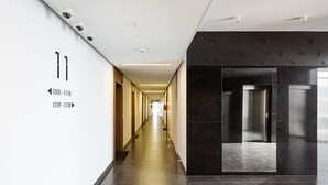 A modern hotel corridor with directional signs, a central elevator, and a reflective black wall. - Wola Chic Luxury Studio (Warszawa)