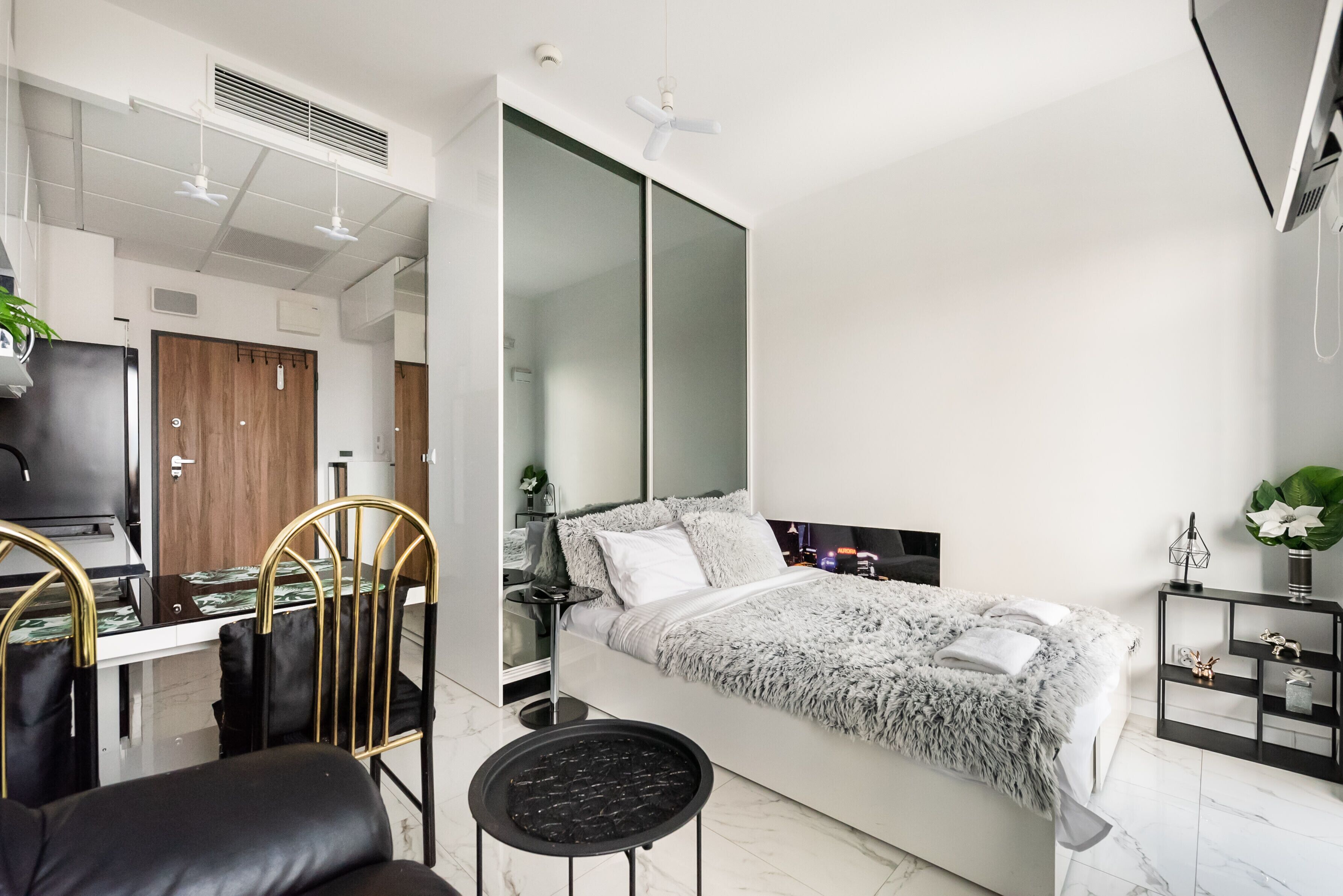 A modern bedroom with a bed, a desk, and a chair. There is a mirror, a ceiling fan, and a small table.