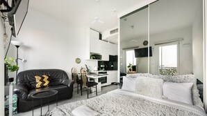 A modern bedroom with a large bed, a black leather sofa, a desk with chairs, and a kitchen area in the background. - Wola Chic Luxury Studio (Warszawa)