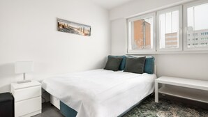 A bedroom with a bed, a bedside table, a lamp, a window with blinds, and a painting on the wall. - Rakowiec Blue Studio (Warszawa)
