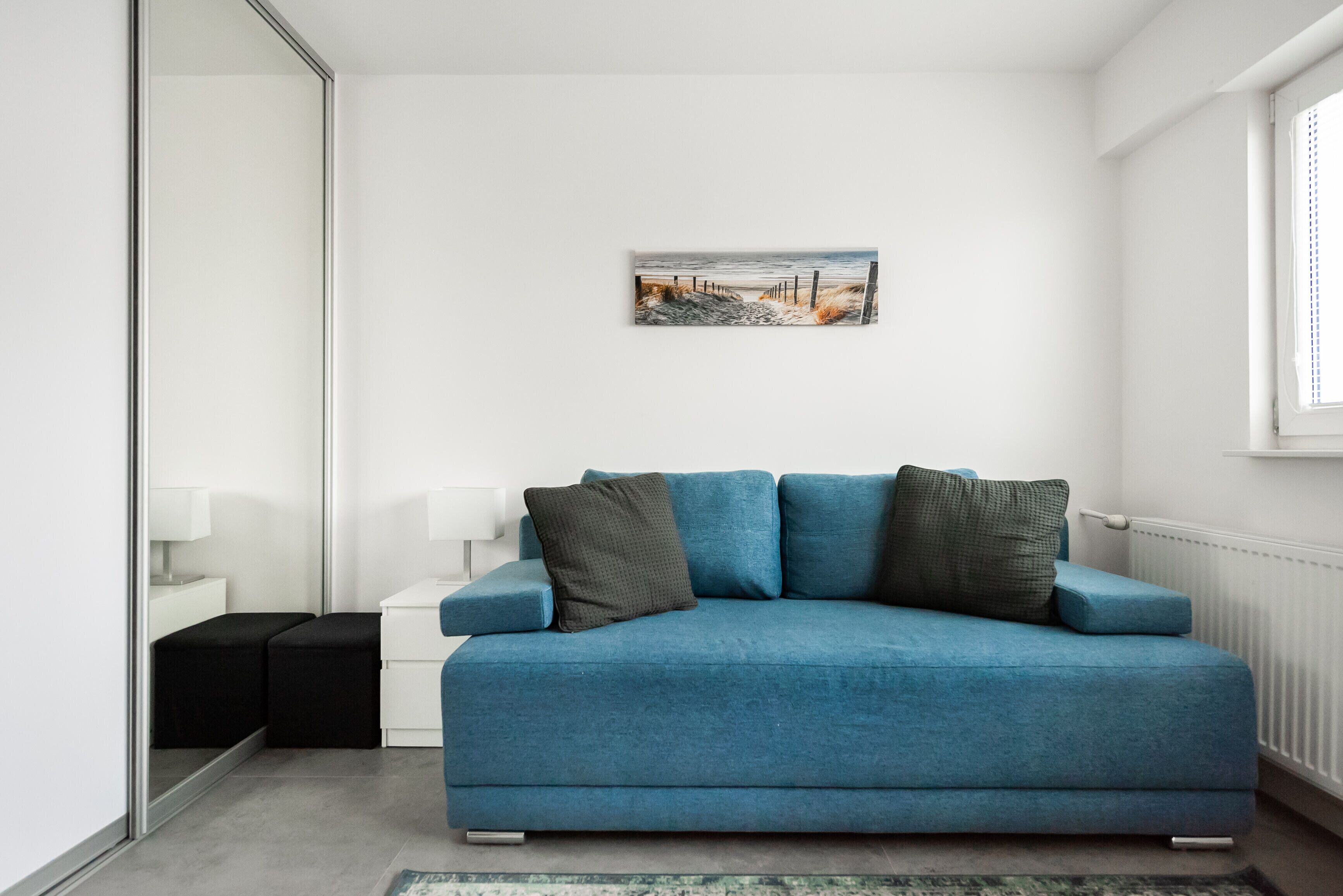 Un salon moderne avec un canapé bleu, une petite table d’appoint blanche, un tableau encadré au mur et une fenêtre avec des stores.