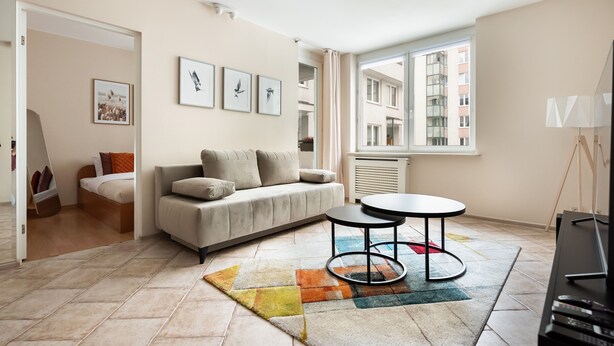 Een moderne woonkamer met een bank, een salontafel en een televisie. Er hangen fotolijstjes aan de muur en een raam met uitzicht op gebouwen.