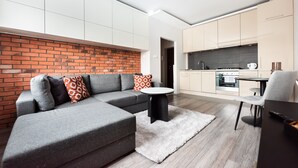 Una sala moderna con un sofá seccional gris, una mesa de centro negra redonda y un tapete blanco. Se ve una cocina al fondo, con armarios de madera clara y un salpicadero gris.