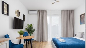 A modern hotel room with a bed, a desk, and chairs. There is a TV on the wall, a window with curtains, and a plant on the desk. - Business Park Blue Studio (Warszawa)
