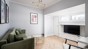 A modern living room with a green sofa, a glass coffee table, a flat-screen TV, and a ceiling light fixture.