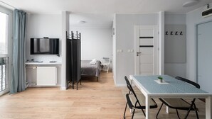 Una habitación moderna y luminosa con una cama, un escritorio con silla, un televisor montado en la pared y una ventana con cortinas.
