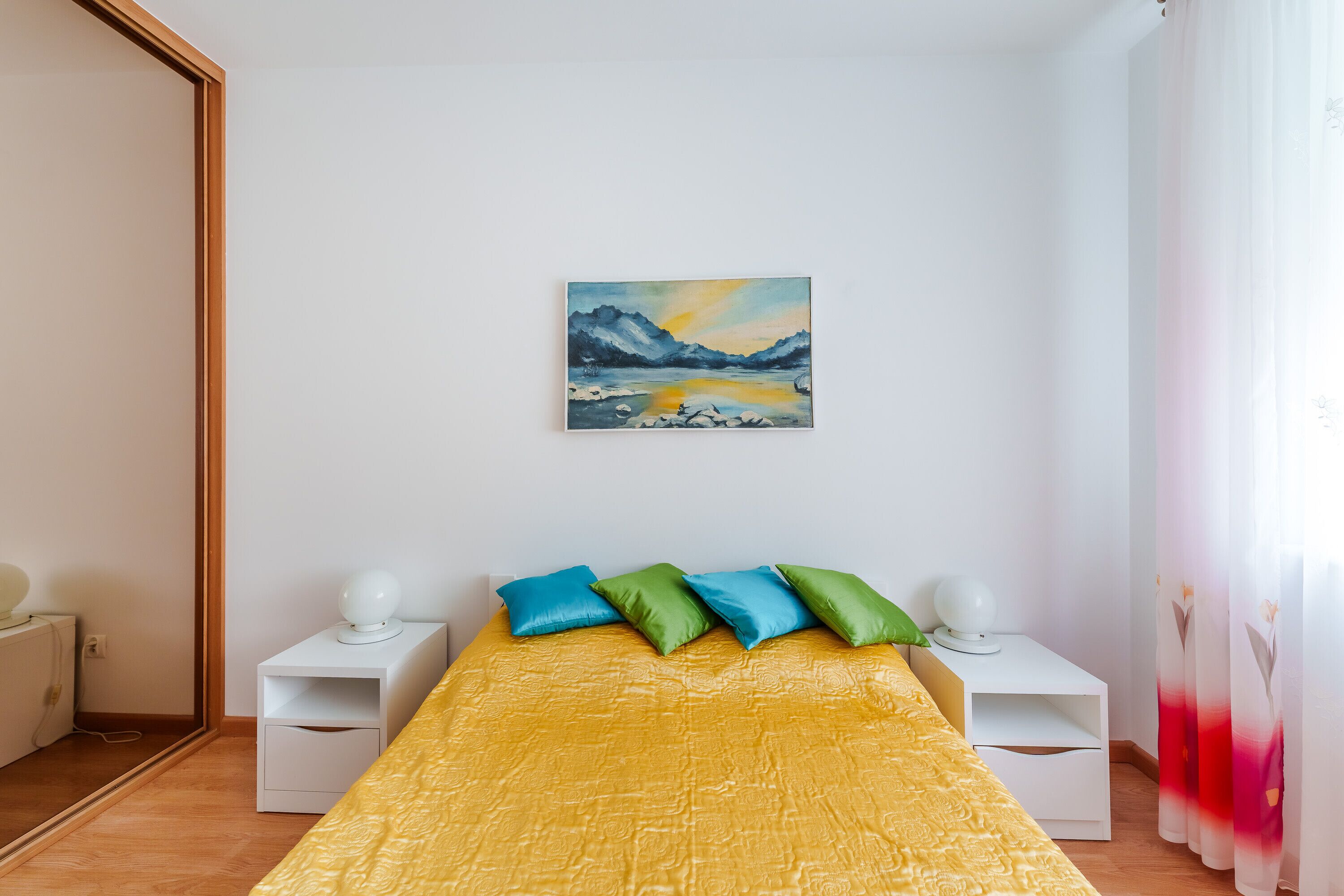A bedroom with a yellow bedspread, a painting of a mountain landscape, and a nightstand with a lamp.