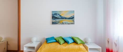 A bedroom with a yellow bedspread, a painting of a mountain landscape, and a nightstand with a lamp.