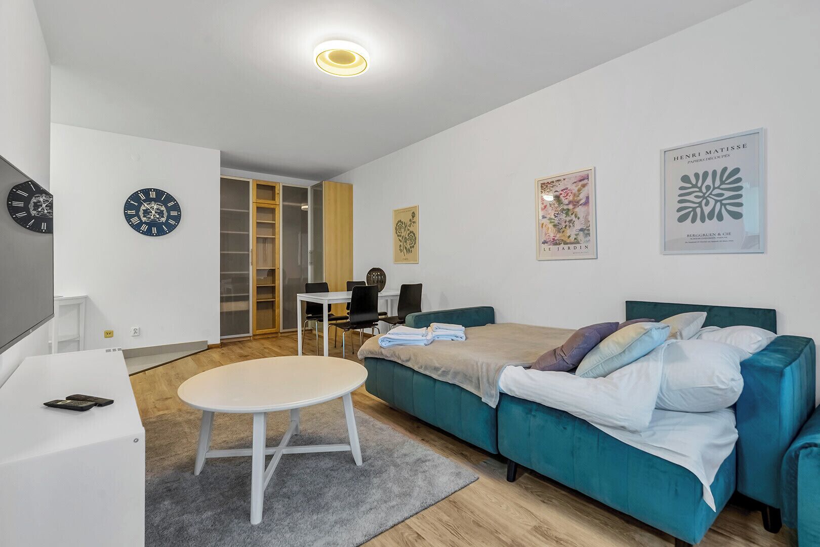 Un salon moderne avec un canapé de couleur turquoise, une table ronde blanche et un téléviseur posé sur un meuble blanc. Deux horloges sont accrochées au mur et une œuvre d’art encadrée se trouve au-dessus du canapé.