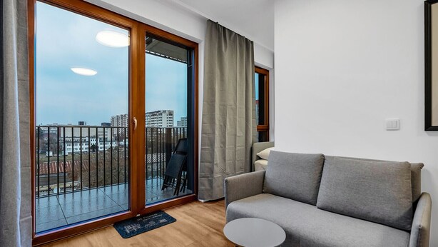 A modern living room with a grey sofa, a small round table, and a view of a cityscape through large sliding glass doors. - Metro Wierzbno Cosy Apartment (Warszawa)