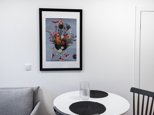 A framed abstract artwork on a white wall, a round table with a glass and two black coasters, a gray sofa, and a black chair.