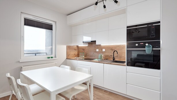 A modern kitchen with white cabinetry, a wooden countertop, a white dining table with chairs, and a window with blinds. - 10th Floor Unii Lubelskiej Apartment (Poznań)
