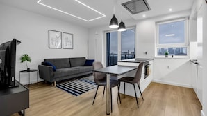 Een modern appartement met een grijze bank, een eettafel met stoelen, een televisie en uitzicht op de stad door grote ramen.