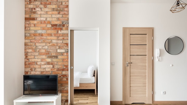 A modern room with a brick wall, a wooden door, a white bench, and a bed in the background.